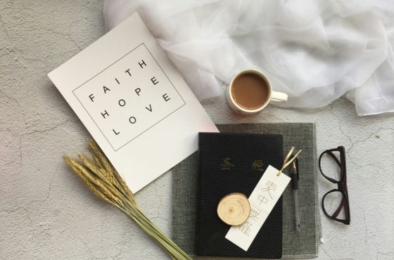 Flat lay of a “Faith Hope Love” notebook, a cup of coffee, eyeglasses, dried wheat stems, and a black journal arranged on a soft textured surface.